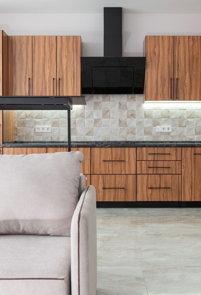 Interior of modern kitchen with wooden cupboards and black exhaust hood placed in room with tiled wall and comfortable sofa